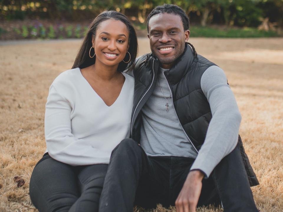 A couple sits in a field and smiles.