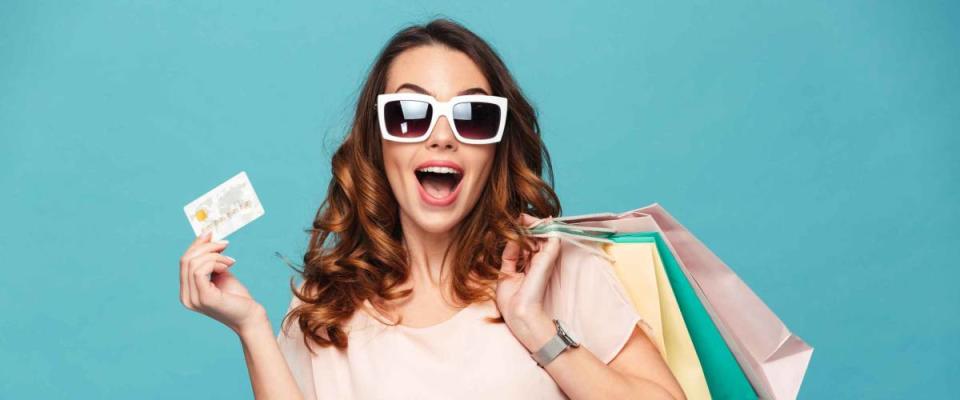 Portrait of a cheery beautiful girl wearing dress and sunglasses holding shopping bags and showing credit card isolated over blue background