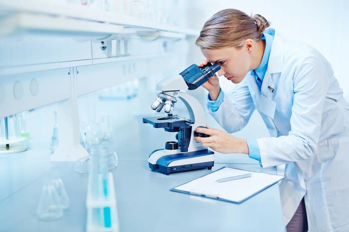 Young blond woman in a white lab coat using a microscope in a lab.