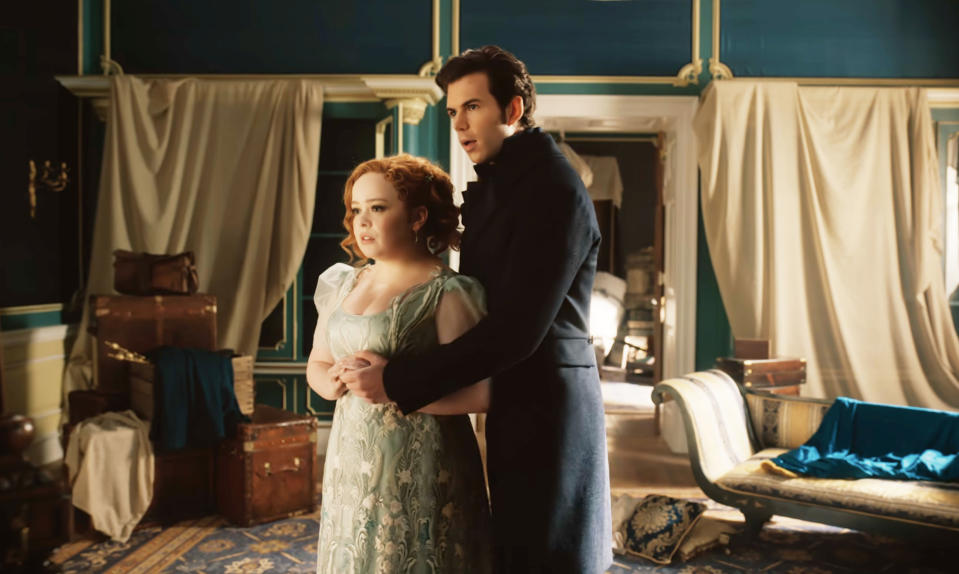 Nicola Coughlan and Luke Newton in period costumes, standing in a vintage room, portraying characters from the TV show "Bridgerton"