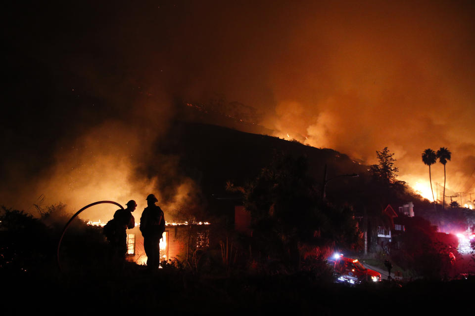 FILE - In this Dec. 7, 2017, file photo, two firefighters watch as a home burns in a wildfire in La Conchita, Calif. Attorneys say as many as 100,000 Californians are eligible to receive payments for the damages they've suffered from a series of devastating wildfires over the last several years, but tens of thousands of them have not. They face a Monday, Oct. 21, 2019, deadline to file claims against Pacific Gas & Electric, the utility blamed for many of the fires. (AP Photo/Jae C. Hong, File)