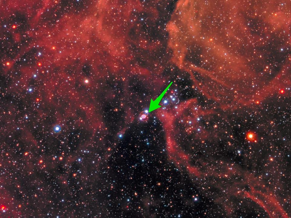 red dusty cloud full of stars in space with a green arrow pointing to a small supernova, which looks like a pink oval-shaped ring with a dot in the center