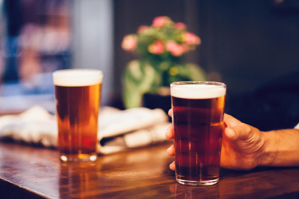 Two pints of beer on a wooden table, with one hand holding a glass. A blurred potted plant with pink flowers is in the background