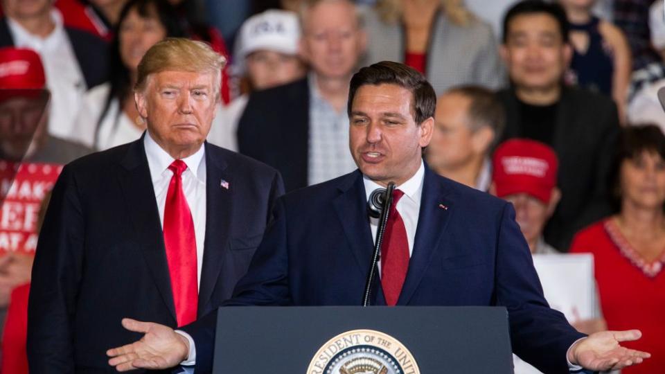 donald trump stands on a stage behind ron desantis who speaks at a lecturn, both men wear suits with white collared shirts and red ties