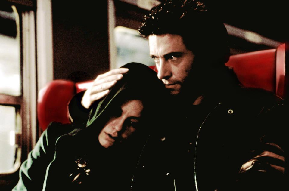 Wolverine in a leather jacket comforts Rogue, who is resting her head on his shoulder, on a train with red seats, scene