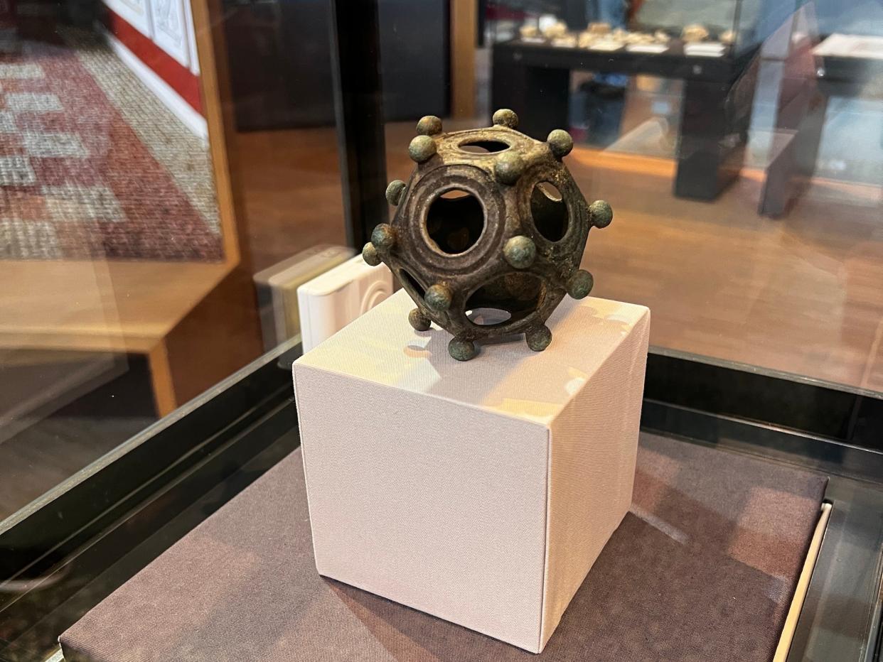A metal dodecahedron with circles on each of its 12 sides and knobs covering it on display in glass case