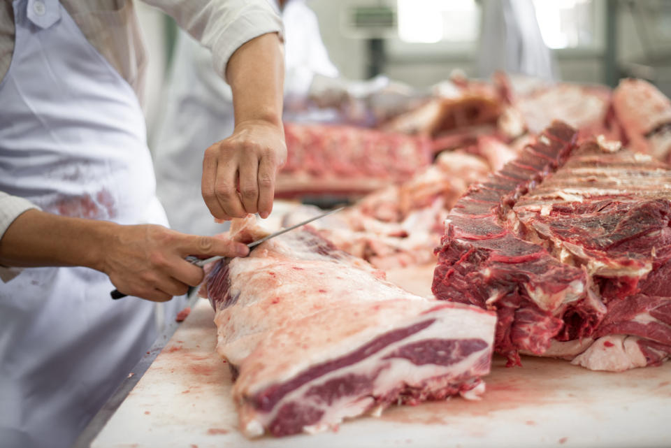A butcher is cutting large pieces of raw meat on a table in a busy butchery. Several pieces of meat and bones are visible