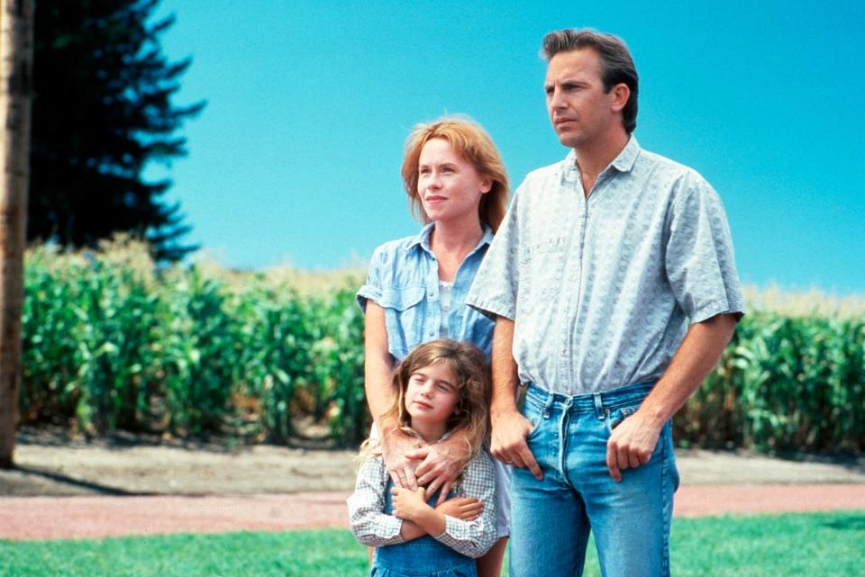 Kevin Costner, Amy Madigan, and Gaby Hoffmann stand together outdoors, with a field in the background, in a scene from the movie "Field of Dreams."