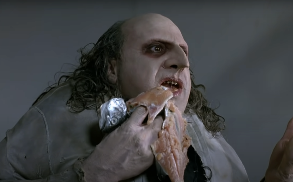 Character portrayed by Danny DeVito in "Batman Returns" grips a fish with his mouth, wearing a stained shirt and coat with frilled edges