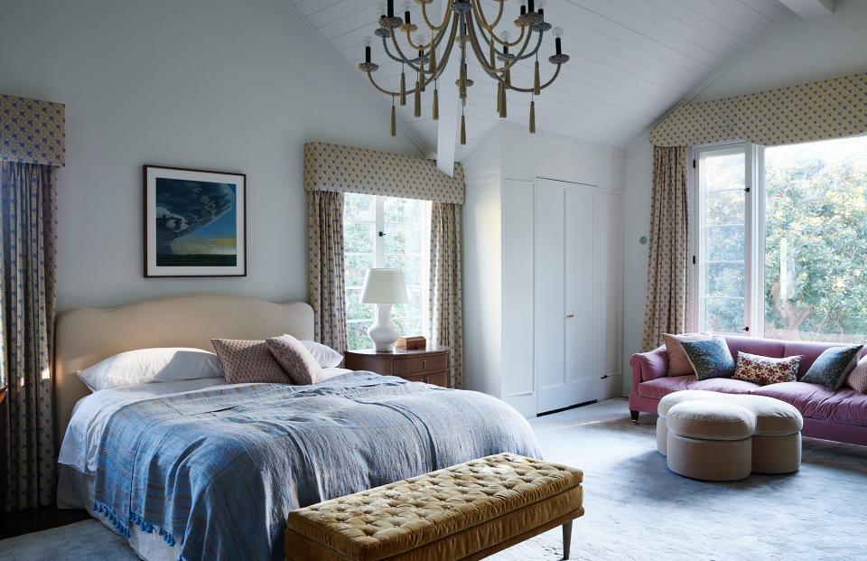 The master bedroom was one of the spaces that underwent the most massive facelift. Levine opened up the ceilings to make them cathedral-style and expanded the windows, making the space airy and bright. The chandelier is by Helene Aumont and sourced from Una Malan, with curtains by Jane Shelton and carpet by Marc Phillips. The couch is custom with velvet fabric from Claremont, the pouf is George Smith, and the bench is from Una Malan. The painting above the bed is Clay Wagstaff from the Sears Peyton Gallery.