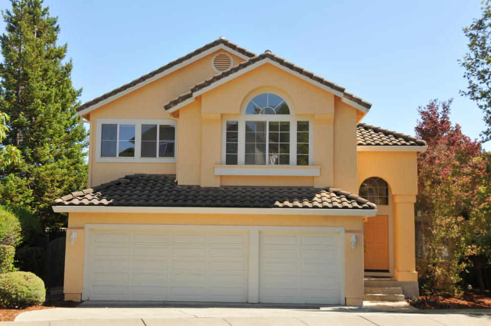 A two-story suburban house with a tile roof, two garage doors, and an arched window above the entryway. Trees and shrubs surround the property