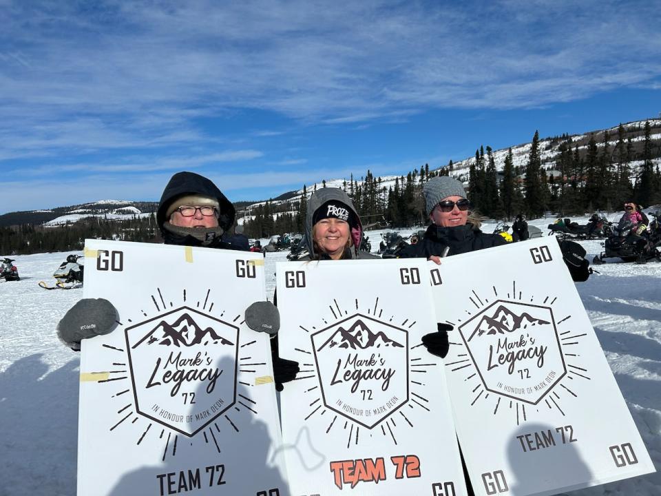 From left: Gen Layvers, Shirley Mood and Kenda d'Eon Amiro stand at the Cain's Quest starting line hold signs cheering on Team 72. The team is racing in honour of Mood's son-in-law, Mark d'Eon.