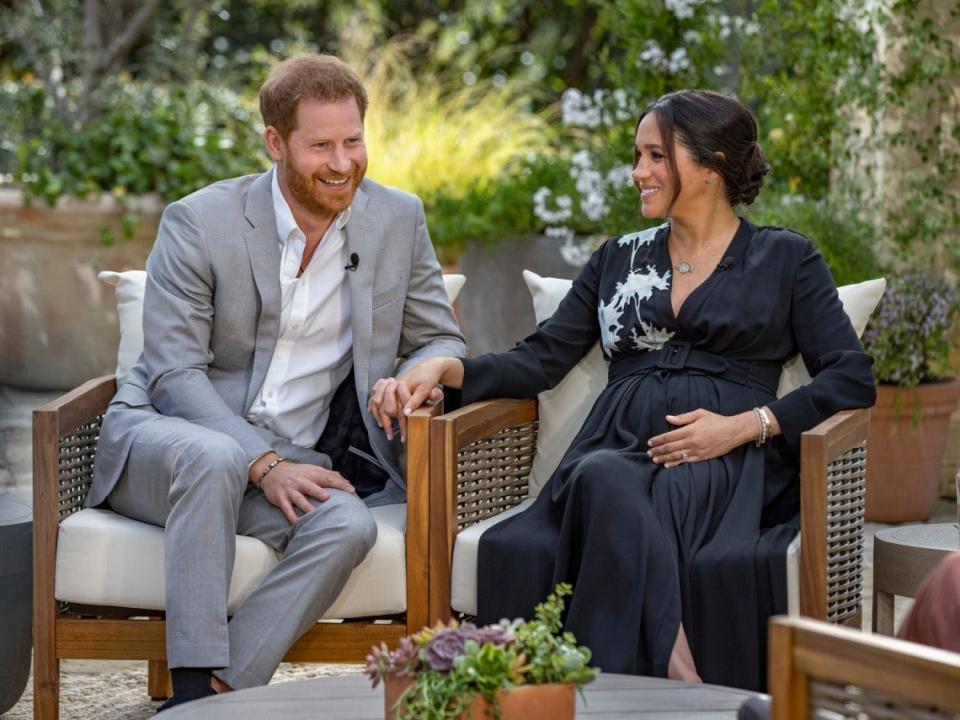 Harry and Meghan smiling and sitting outside and holding hands in formal attire.