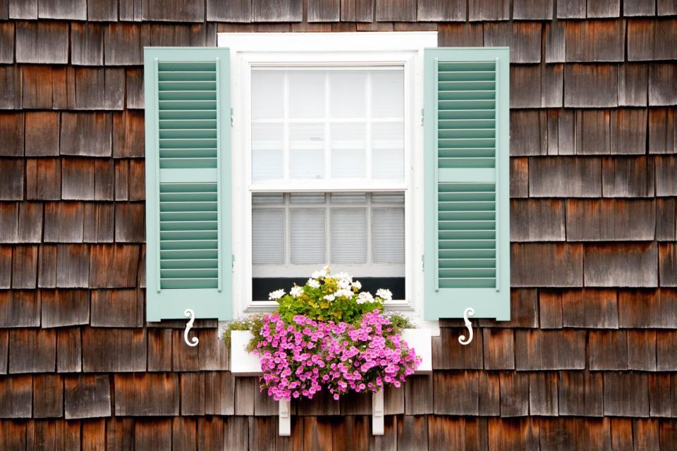 Old beach cottage with wood shake siding. Window with turquoise shutters and white wooden window box and trailing pink and white flowers in summer.