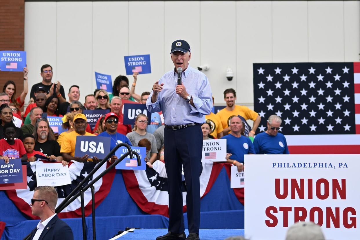 Biden points his finger and wears his presidential seal hat and holds a mic on a stage in front of an American flag and next to a sign that says "Union Strong."