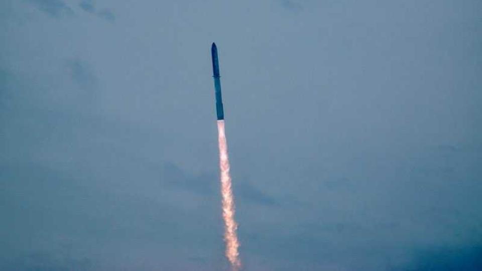 a large silver rocket flies through a blue sky above a huge column of fire