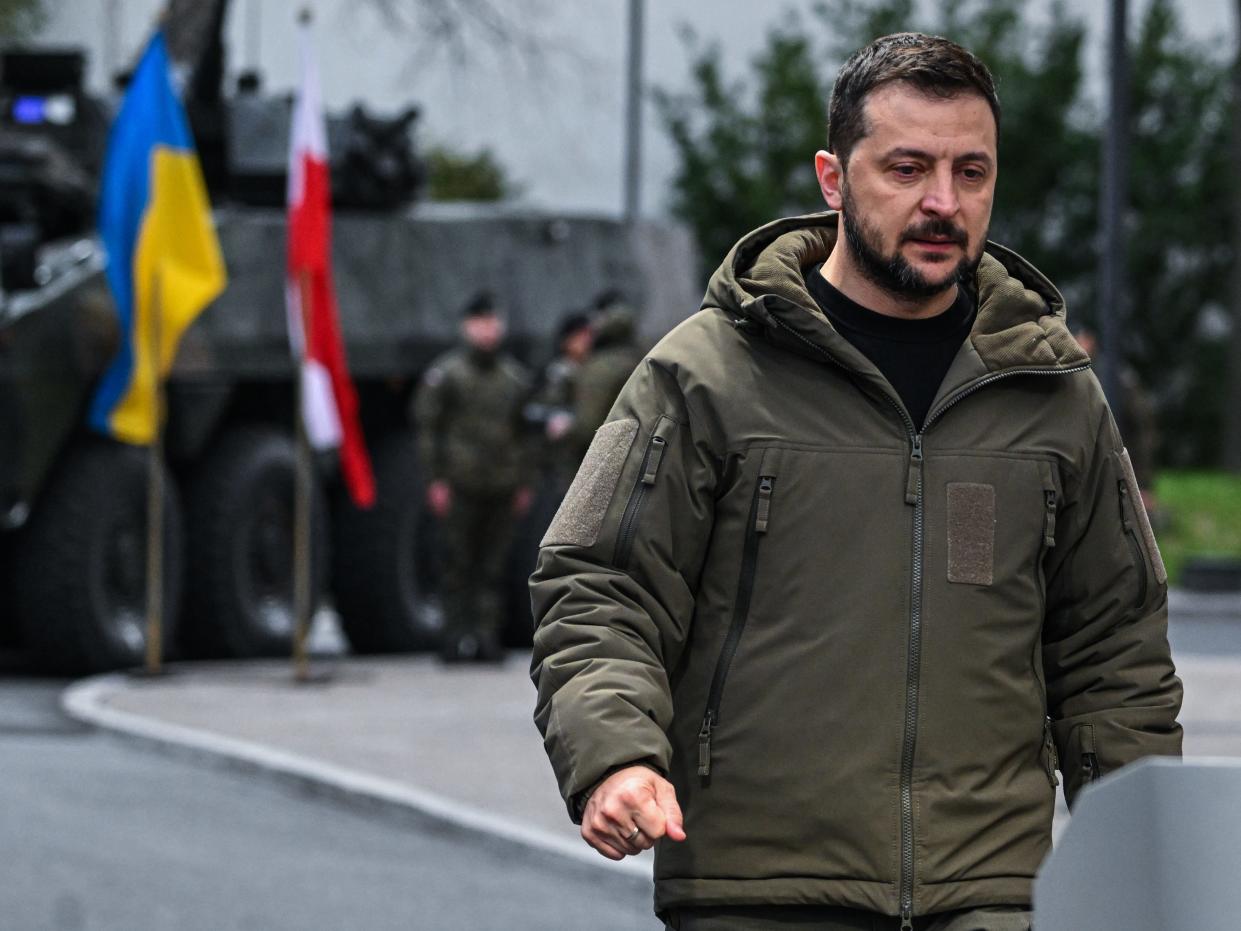 Zelenskyy in coat, armored vehicle and Ukrainian and Polish flags in the distance behind him