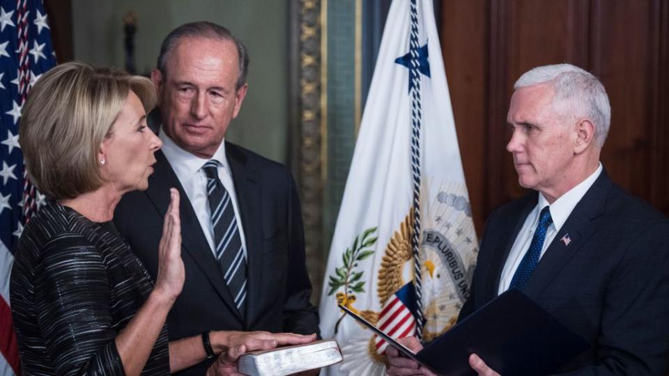 betsy devos stands with one hand raised and the other on a book held by husband dick devos, she looks at and speaks to mike pence who holds an open folder, behind them are two flags