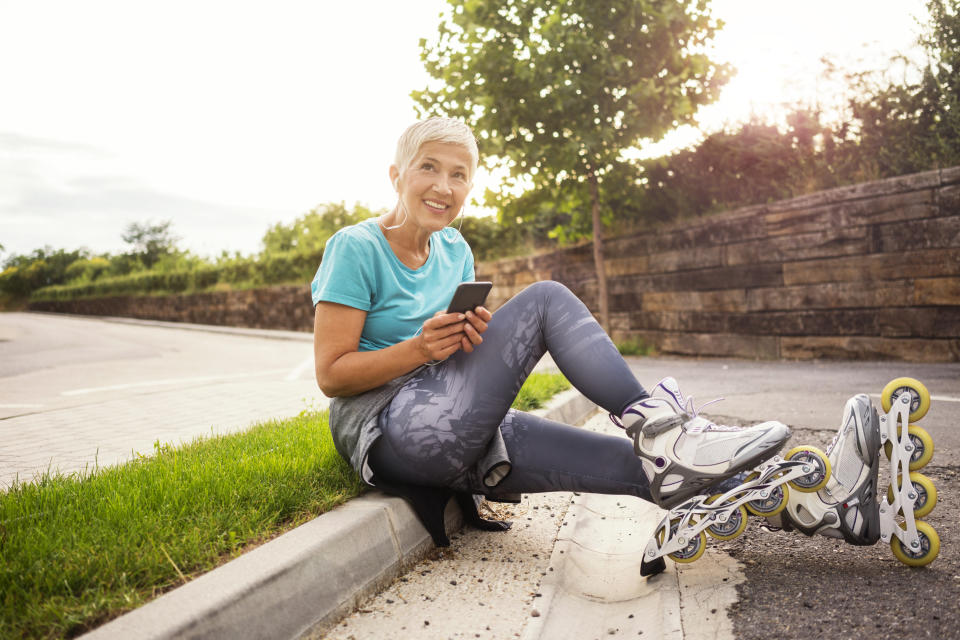 An older woman sits on a curb holding a phone with inline skates on her feet, smiling at the camera