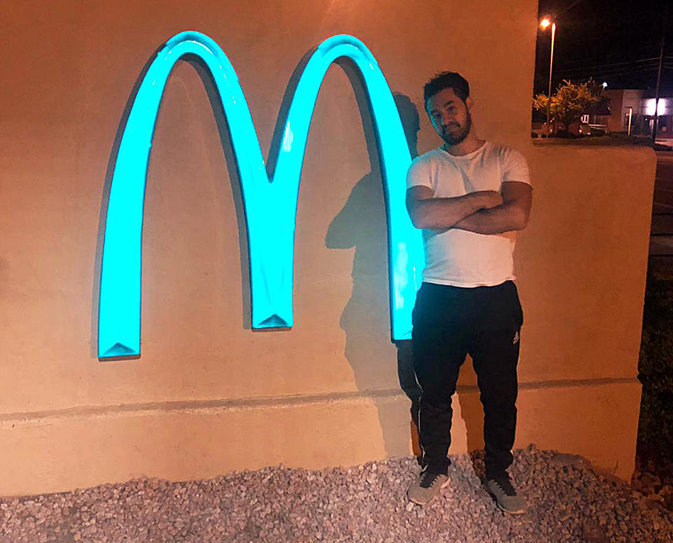 In this 2018 photo provided by Regina de Heer, Joseph Gedeon poses next to the arches of a McDonald's restaurant, which are among the very few in the world that are not golden, and the only one in the world that are turquoise, in Sedona, Ariz. To preserve its beauty, the city of just over 10,000 people has a strict building code and zoning laws: Structures can’t grow too high, and must be colored in hues that complement the natural tones of the red rocks. The arches are turquoise to enhance the desert’s natural beauty. (Regina de Heer via AP)