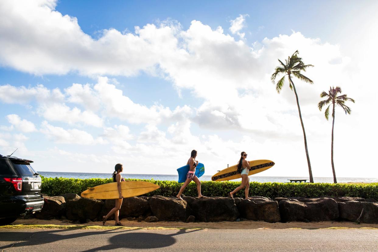 Three people hold surf boards and walk towards the ocean. Their car is parked behind them, and the blue sky and tons of clouds are overhead. Two palm trees are also in front of them.