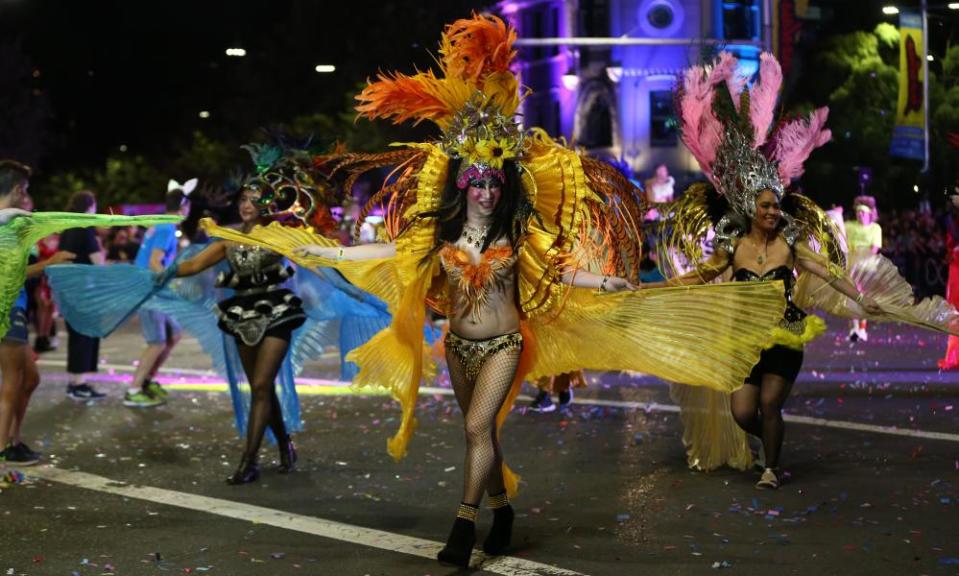 A drag queen in flame orange with a large feathered head-dress and a pleated chiffon cloak that looks like wings ruffles her costume in front of a group of other dancers, in similar outfits, in the middle of the road at night during Mardi Gras.