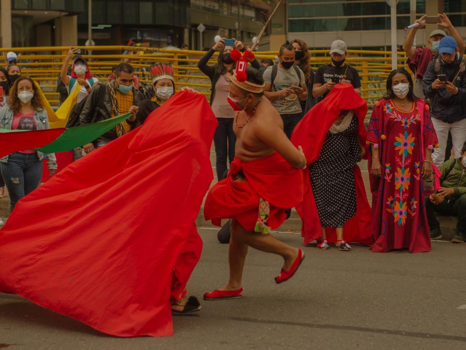 A shirtless man with red fabric draped around him dances in the street with a crowd of people watching