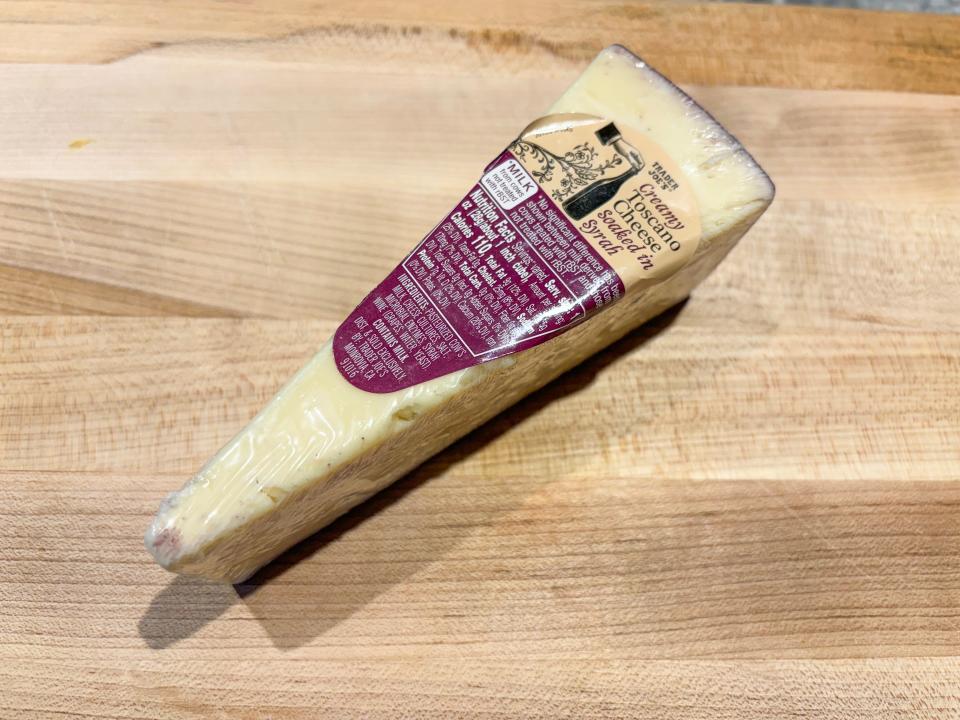 A yellow wedge of cheese with a red rind and a purple and peach label reading "creamy Toscano cheese soaked in Syrah"