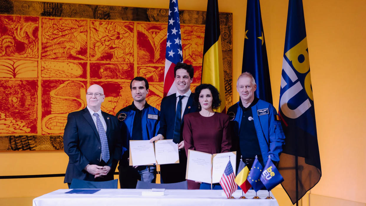  Five well-dresse dpeople stand in front of furled flags in a yellow-walled room in front of a white table, which holds several small flags. 