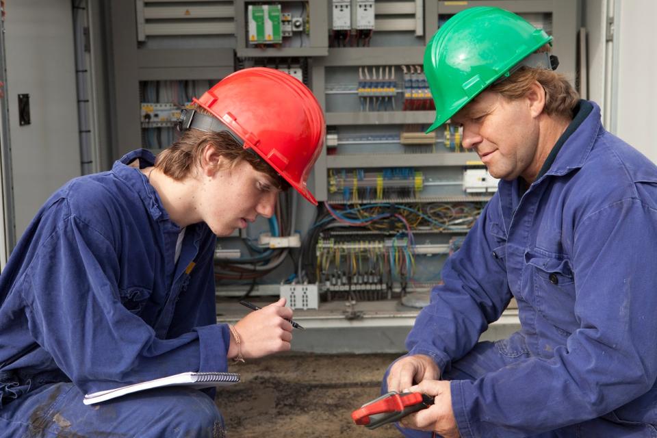 Two electricians in blue jumpsuits examine a large electrical board. 