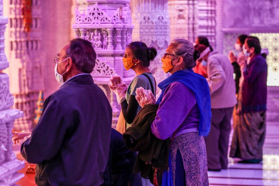 People wearing masks and plastic gloves stand inside a Hindu temple with their hands pressed together in prayer