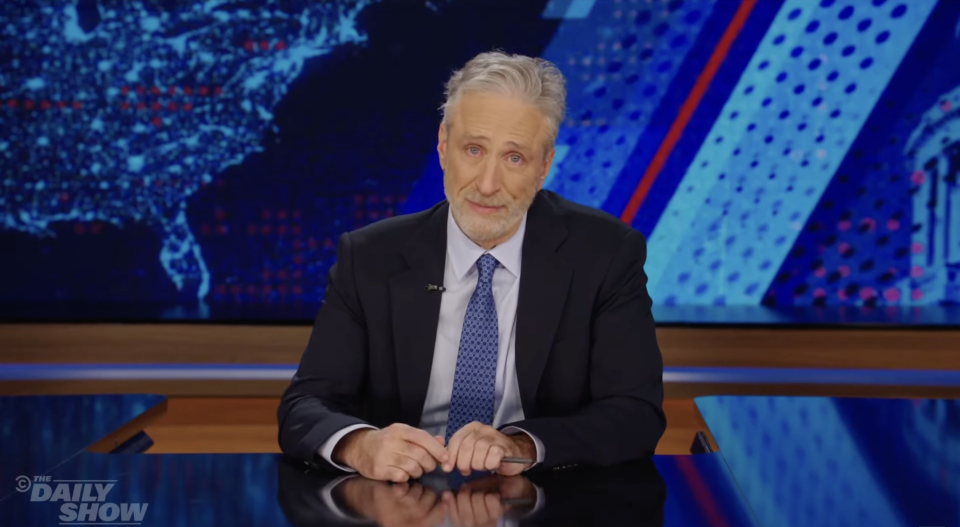 Jon Stewart in a suit hosts "The Daily Show," sitting at a desk with the show's graphics in the background