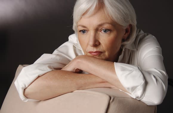 A worried senior woman with her arms crossed and propped on the back of a chair, and her chin resting on her forearms.