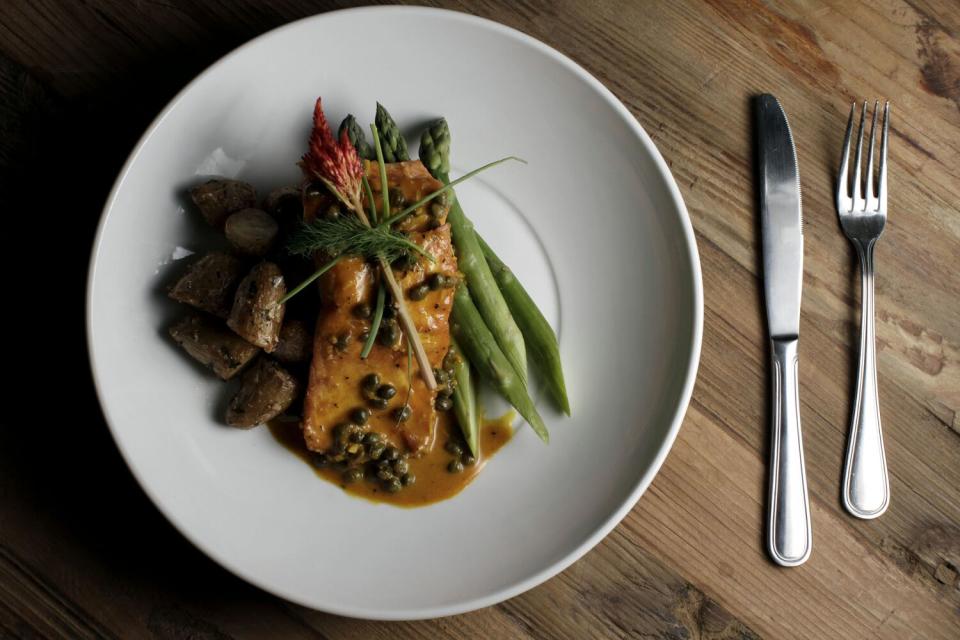 A plate with salmon, capers, asparagus and potatoes sits next to silverware.