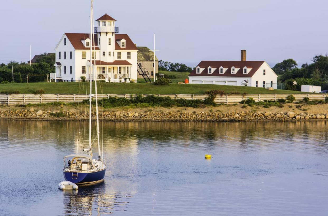 boat on the water with a shore with a white picket fence and two big white buildings with a red roofs behind it