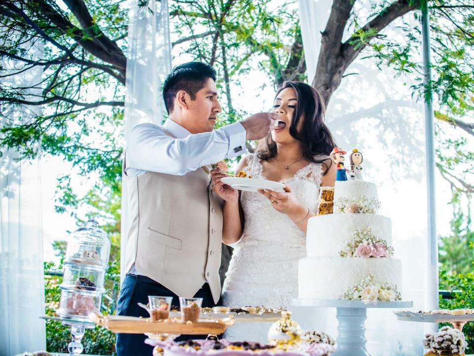 A groom feeds a bride a bit of cake in front of trees. Their cake with a "Mario"-themed topper sits on a table in front of them.