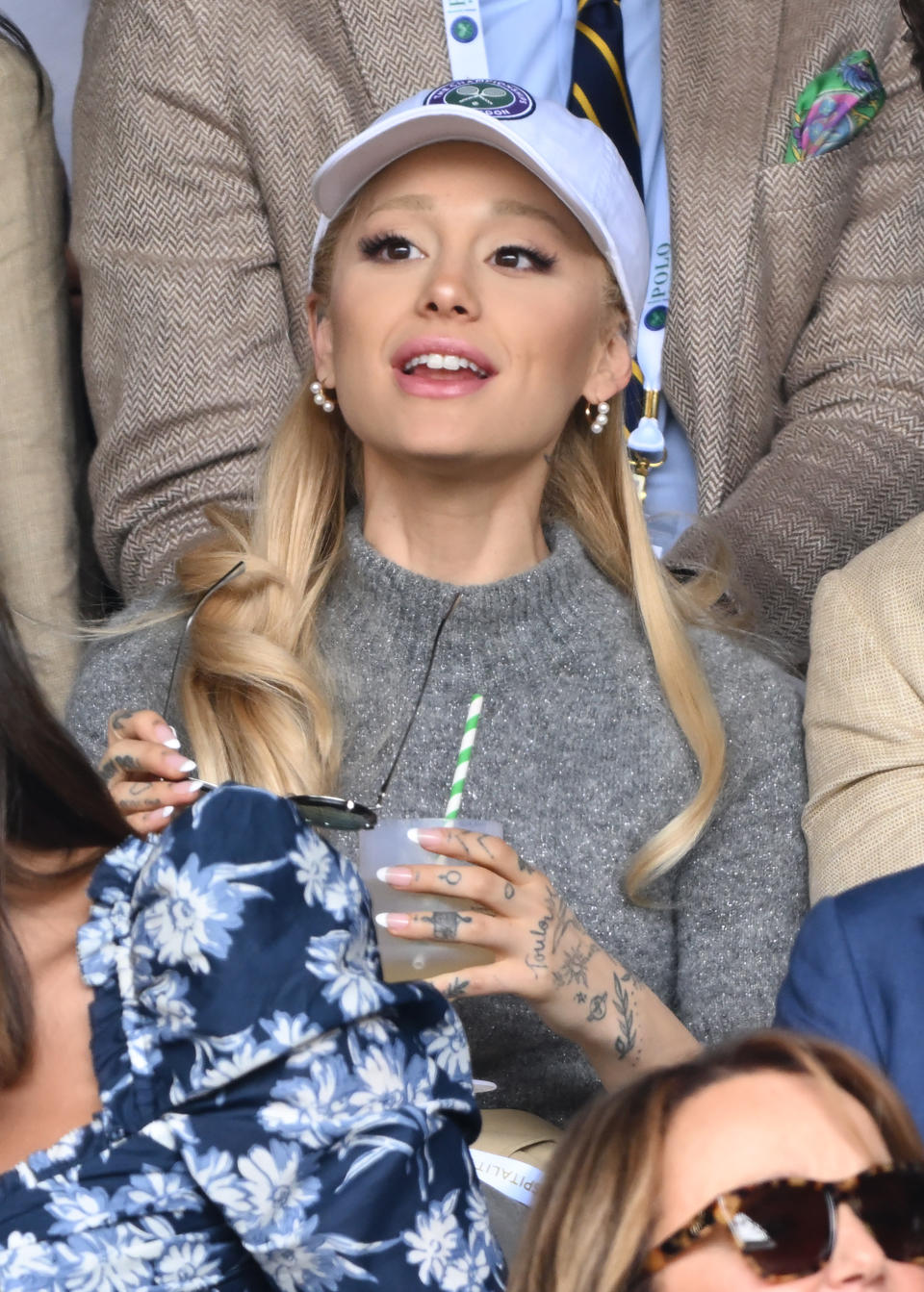 Ariana in a sweater and cap, holding a drink & watching a tennis match