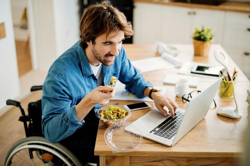 young businessman in wheelchair eating while surfing the net on laptop on lunch break at home