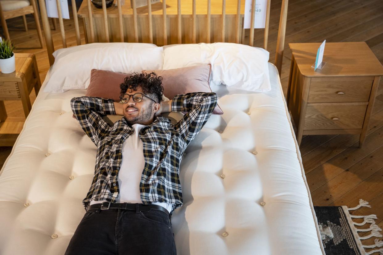 A mid adult man lying on a bed in a furniture store, he is testing it out and considering to buy it. He has his arms behind his head and is looking away from the camera.