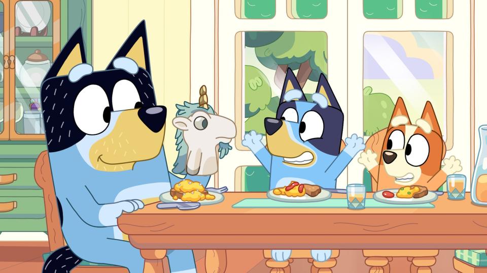 Bandit, Bluey and Bingo Heeler sit at a dining table in the animated show Bluey.