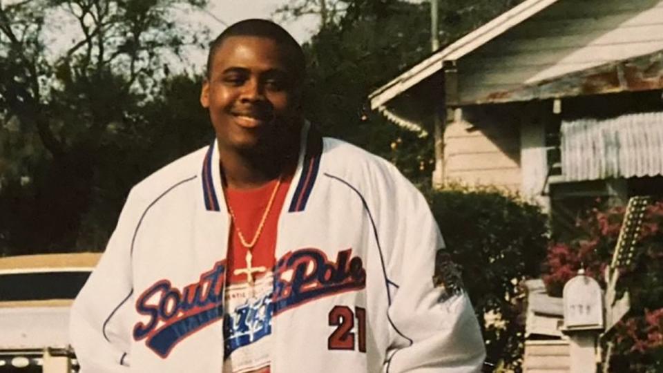 Lashawn Thompson smiling in front of the mailbox of a small house, with a white letter jacket saying South Pole, and a cross around his neck. 