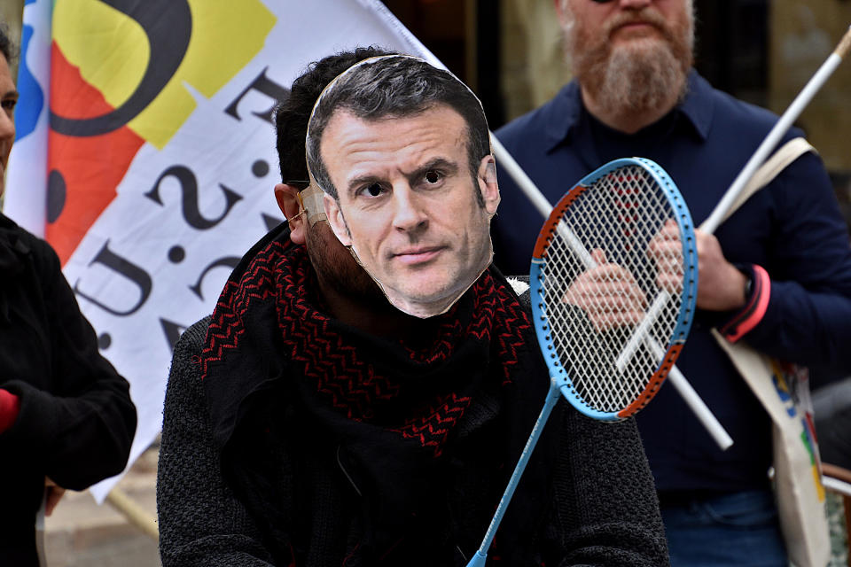 A protester wears a homemade mask with a picture of Macron's face and holds a badminton raquet