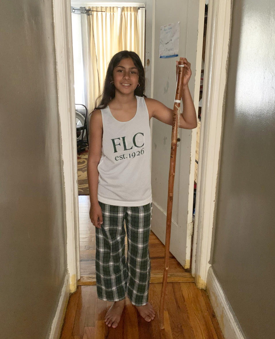 In this June 22, 2020, photo provided by Roxanne Bridglall, 10-year-old Annabelle Bridglall appears at her home in New York, holding her commemorative walking stick from Forest Lake Camp, where she has spent the last three summers. Every summer, the campers get a new mark on their stick to signify how many years they have attended the camp. This is the first time in the camp’s 95-year history that it will not be open due to state coronavirus restrictions. (Roxanne Bridglall via AP)