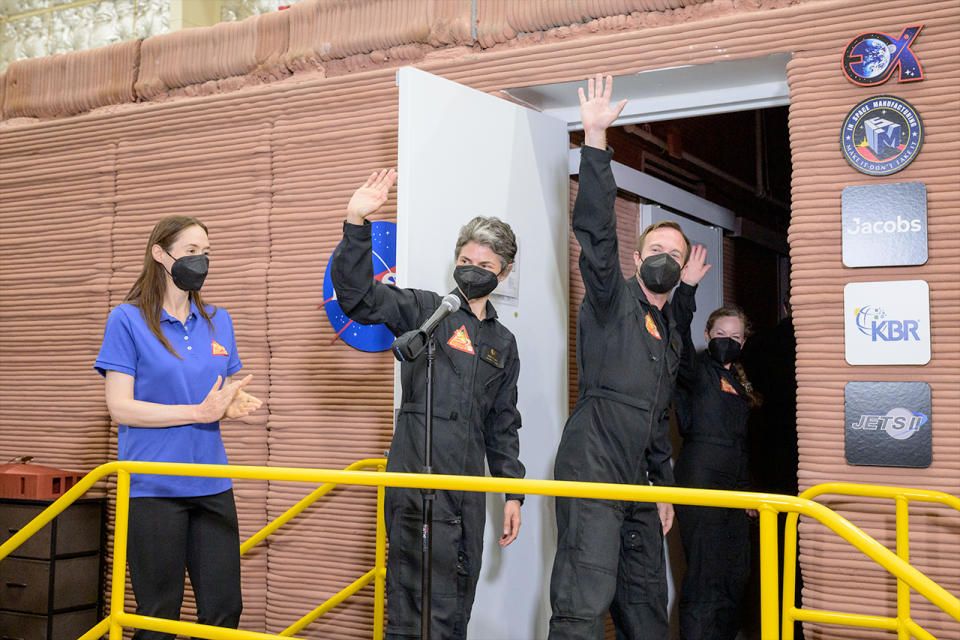 three people in black jumpsuits wave as they enter a brick building