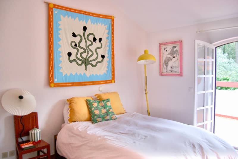 Tapestry artwork hung above bed in white bedroom with wood chair, yellow floor lamp, and French doors.