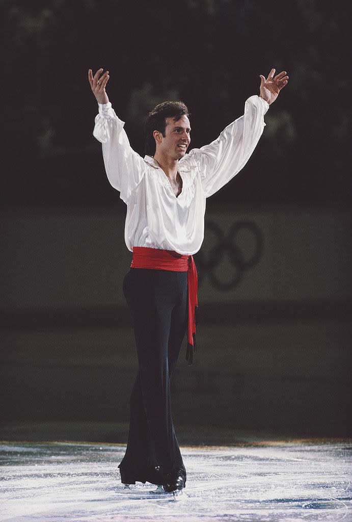 Brian is wearing a large blouse-like shirt as he stands on the ice and raises his arms in acknowledgement of the crowd