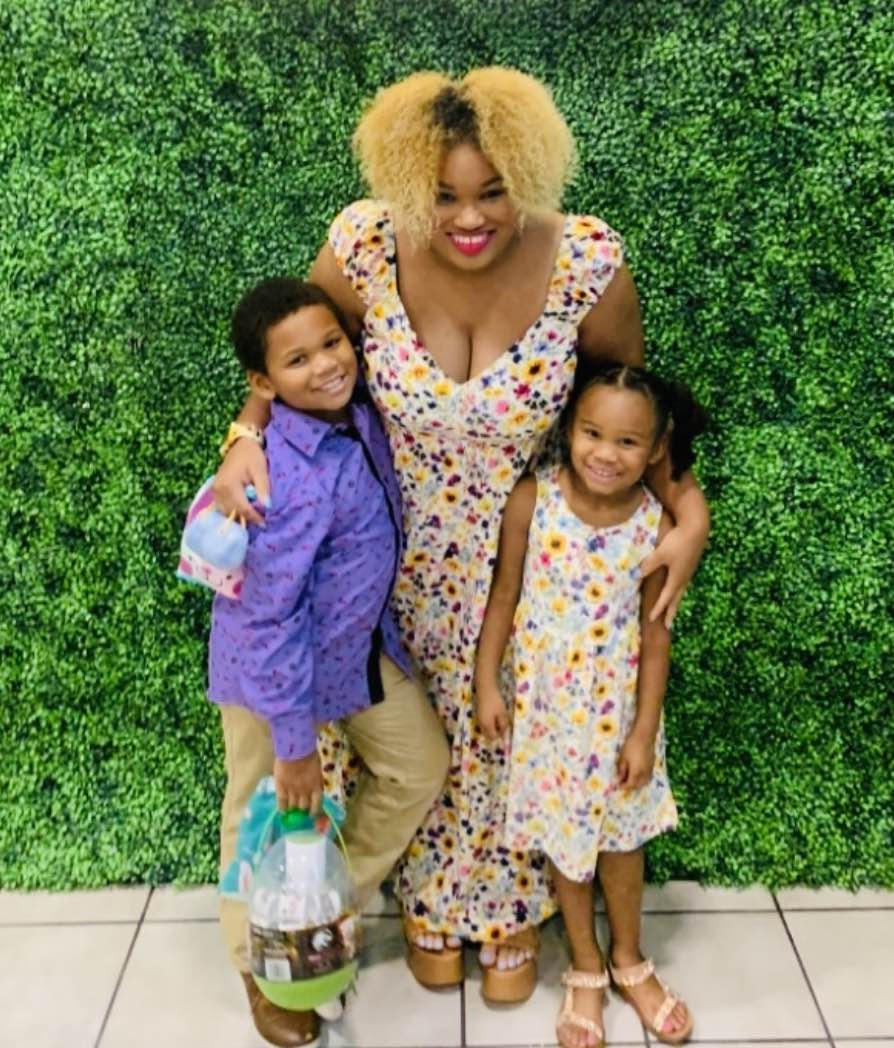 Smiling mom, who has epilepsy, stands next to her two young children in front of a green hedge.