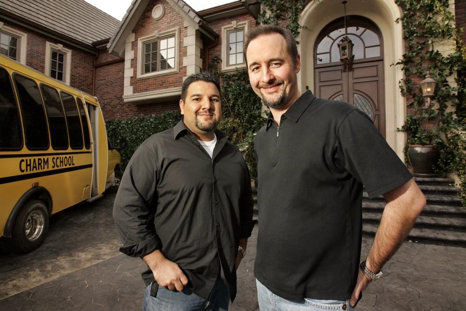 Two men in black shirts smile in front of a mansion and next to a school bus