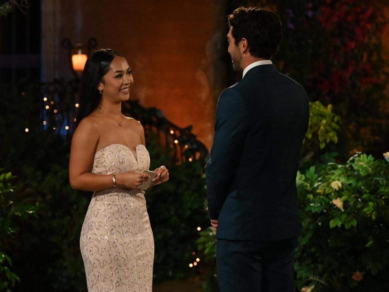 Lea smiles in a strapless gown with gold sequin detailing outside of the Bachelor mansion while Joey, in a blue suit, stands before her.