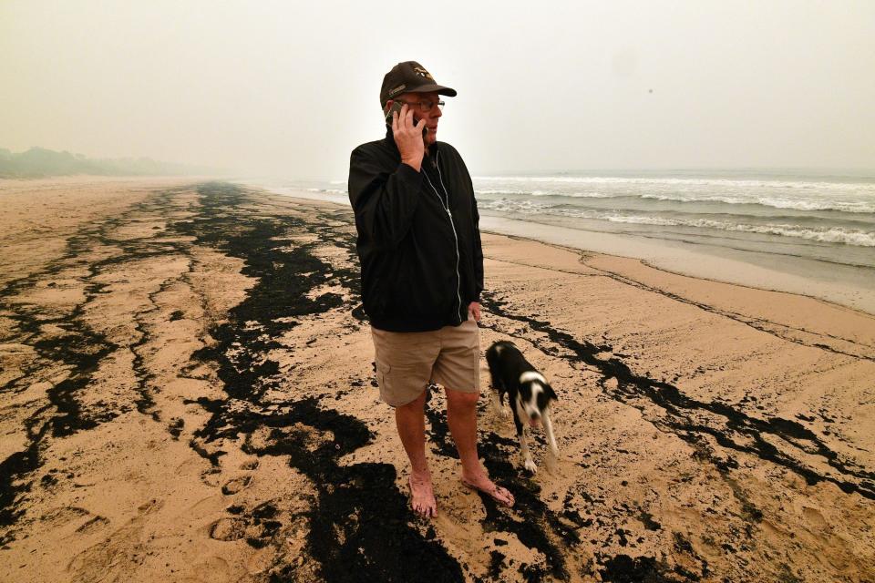 Resident Dave Iredale talks on his phone while standing amongst ash from bushfires washed up on a beach in Merimbula.